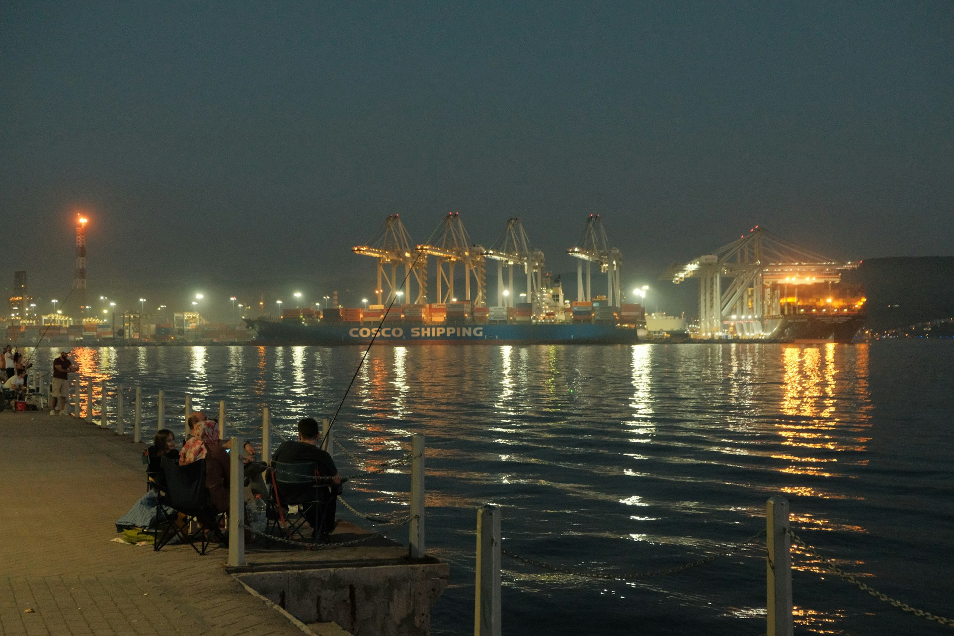 People fishing by a harbor with cargo ships at night.
